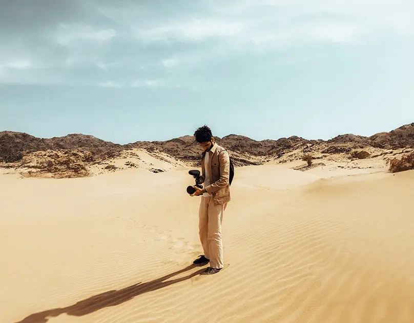 Azzam holding a cinema camera on a desert dune under a pale sky