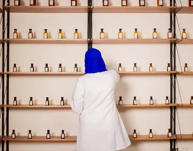 A perfumer in a white coat and blue hijab reaching for a bottle on tall shelves